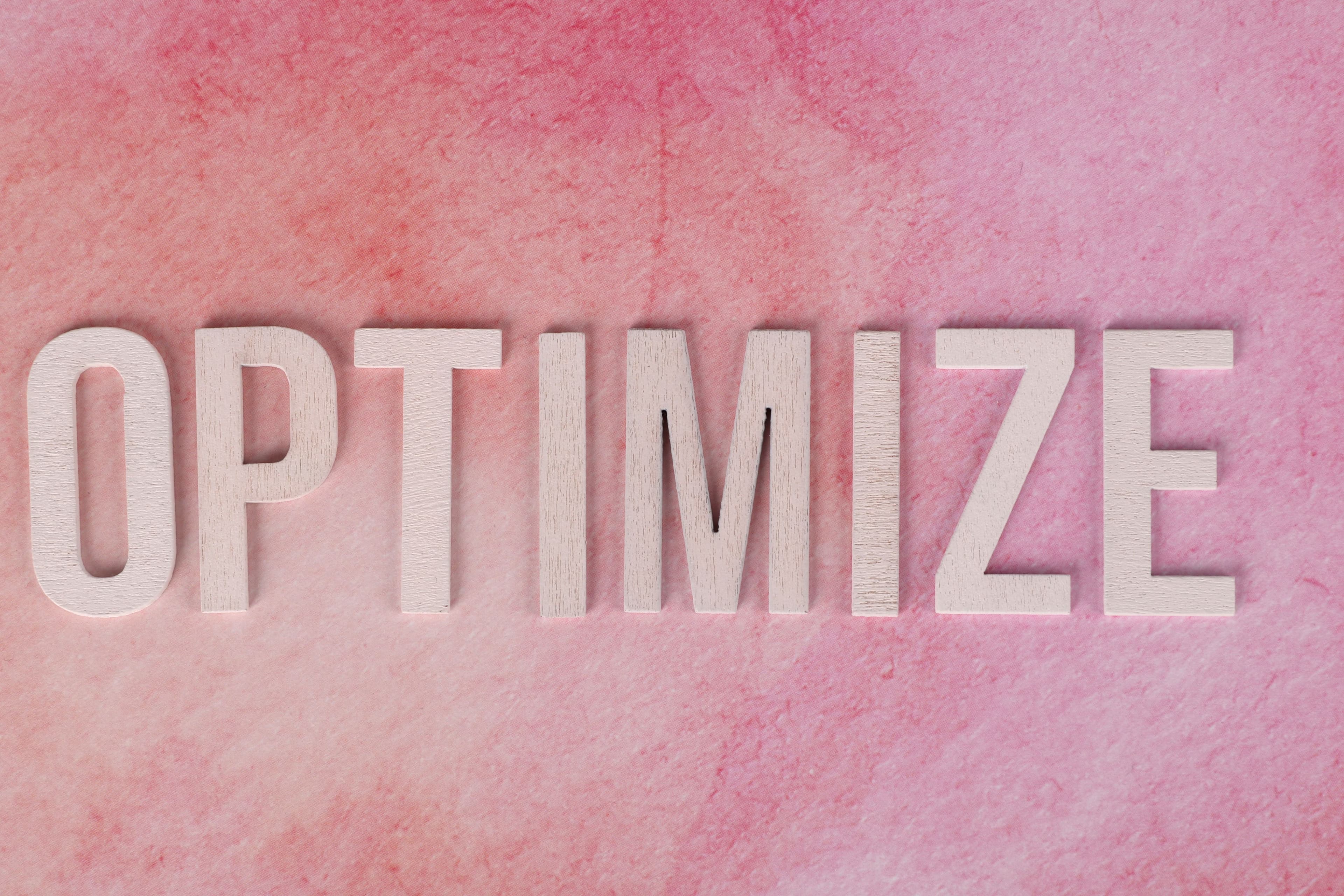 White wooden letters spelling “OPTIMIZE” against a textured pink background.