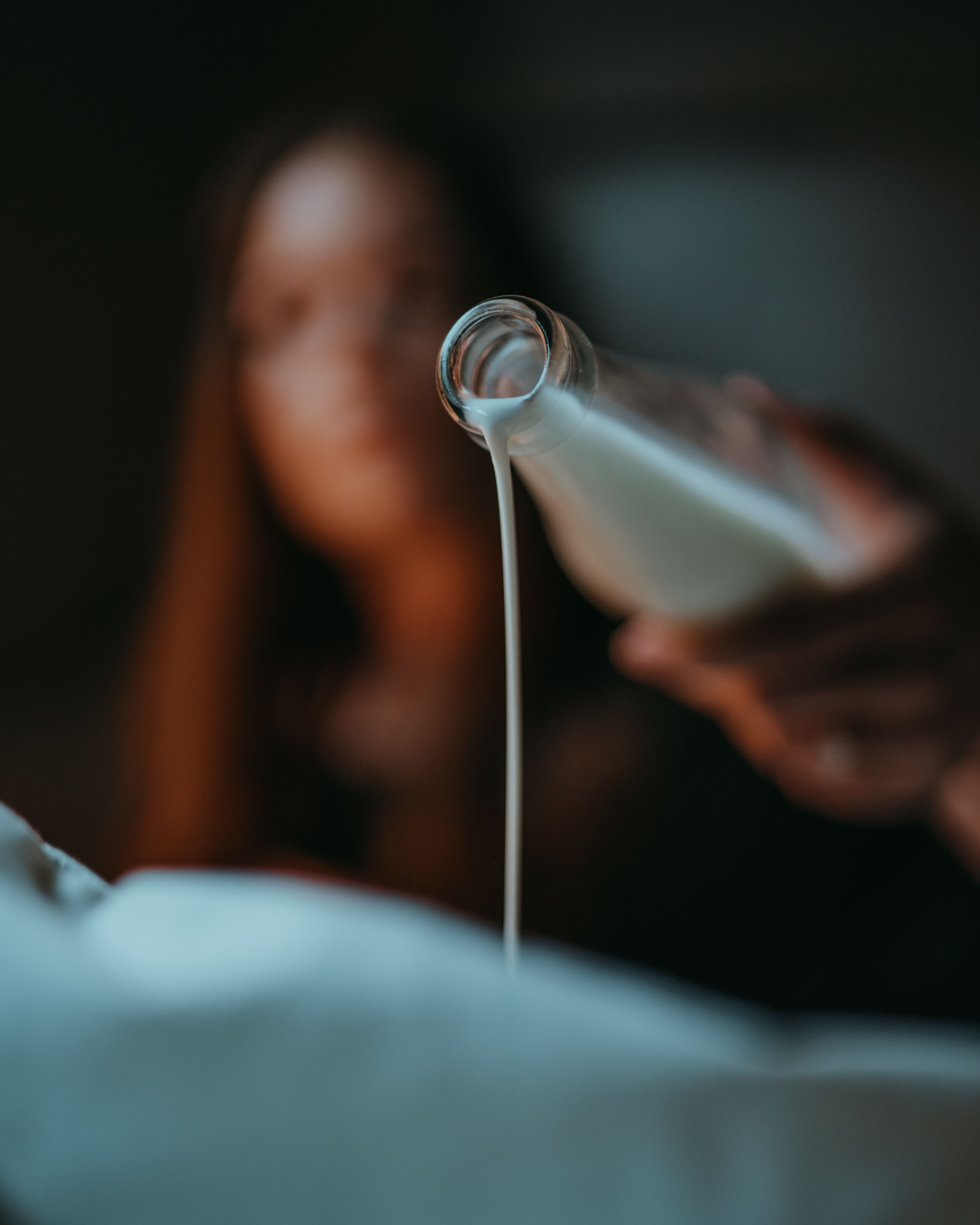Close-up of a person pouring a thin stream of white liquid from a glass bottle onto a surface, with the person blurred in the background.