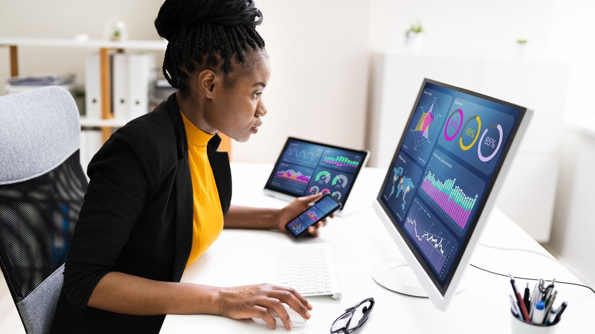 A woman at a desk reviews analytics charts on a monitor, tablet, and phone