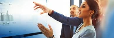 A woman gestures toward charts on a large display while a colleague stands nearby