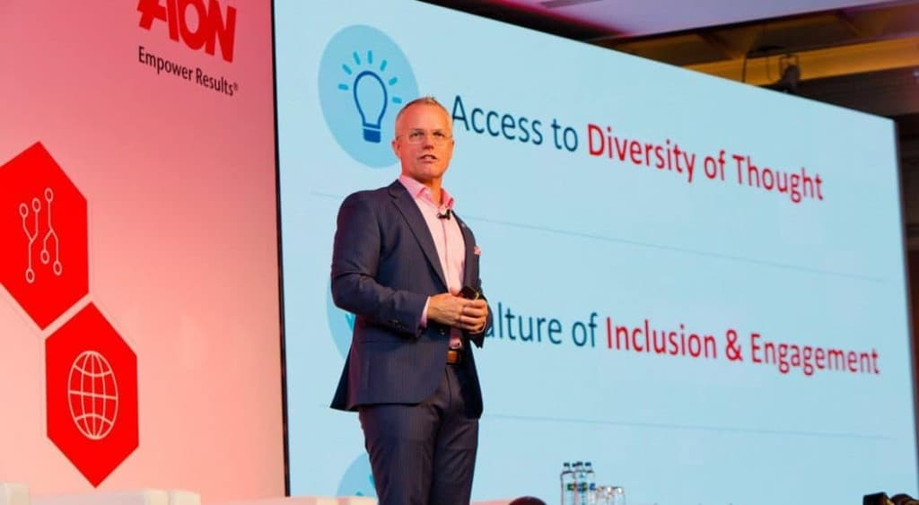 A speaker stands on stage beside a screen that reads Access to Diversity of Thought and Culture of Inclusion and Engagement, with Aon branding on the left