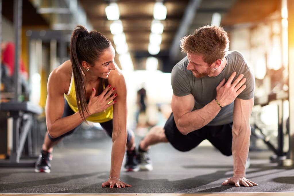 Two people hold a plank in a gym and look at each other.