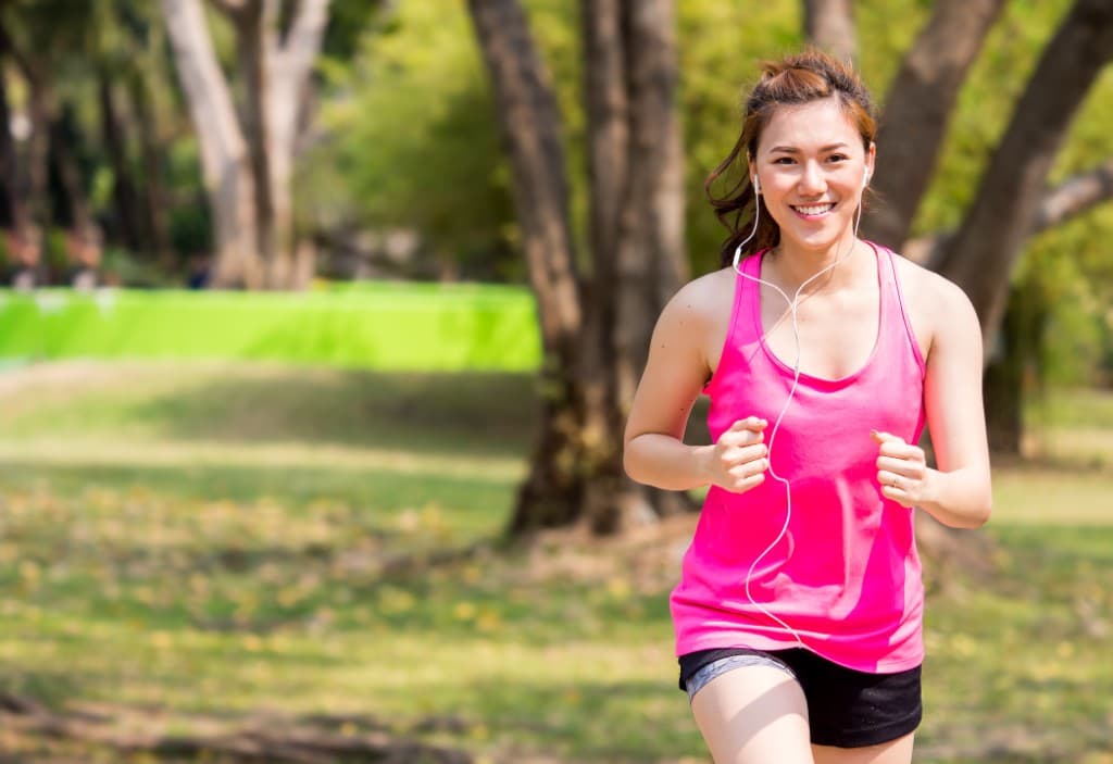 A woman in a pink tank top jogs through a park with earphones in.